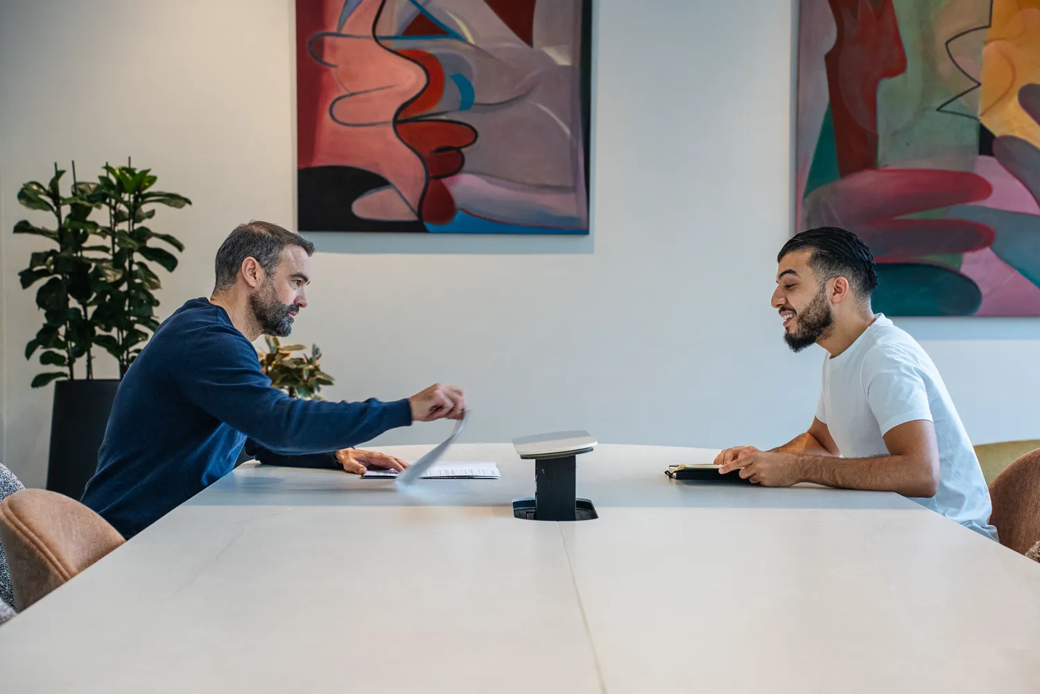 Deux collègues en discussion à une table de travail, plantes et tableaux en arrière-plan